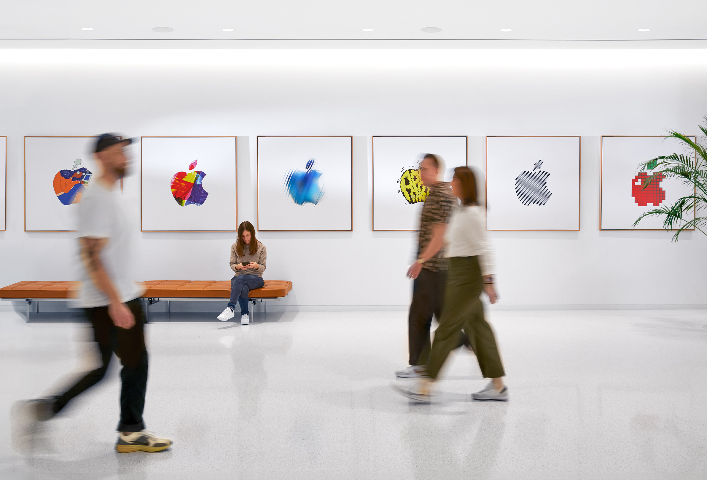 Tři zaměstnanci Apple procházejí v interiéru kolem stěny s barevnými logy Apple a jedna zaměstnankyně sedí na lavičce.