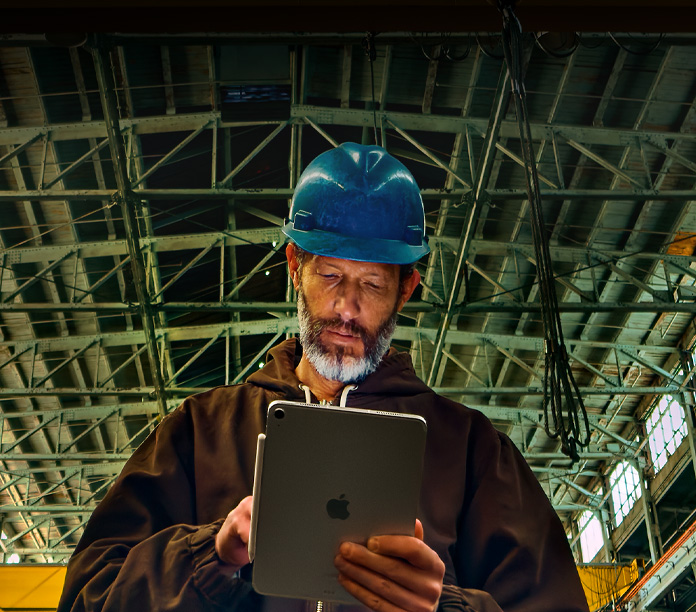 iPad, silver, with Apple pencil attached held by a person in a brown jacket and blue hard hat, working in an industrial setting with metal beams overhead