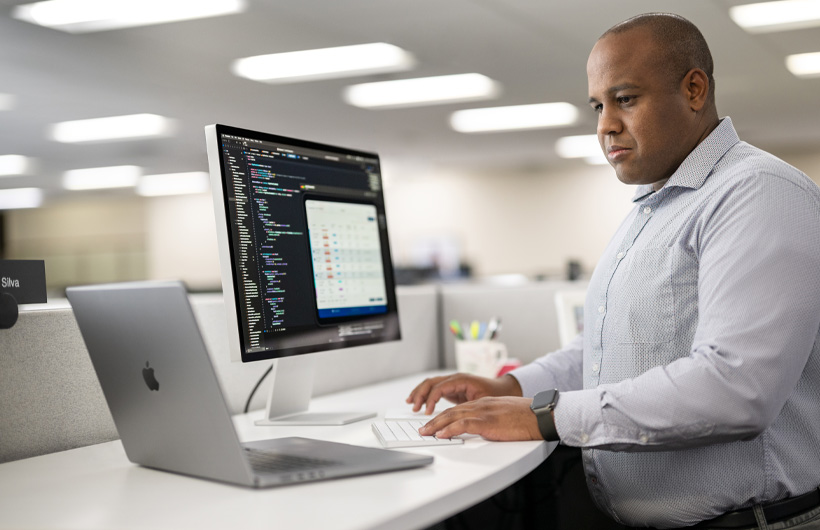 Programador sentado à secretária a usar um Mac e um MacBook, com o ecrã do Mac a mostrar código e informações da app