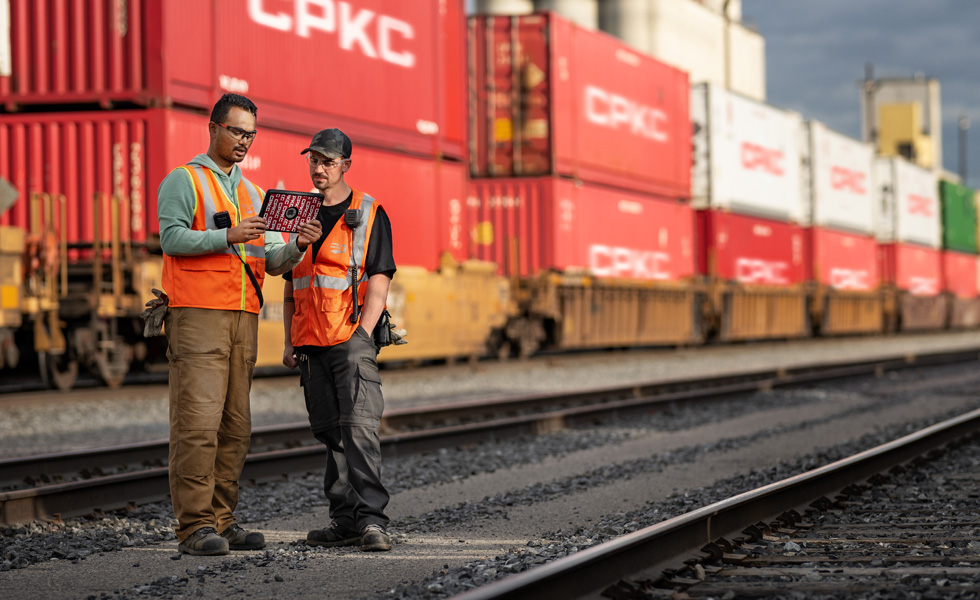 Dois trabalhadores ferroviários a olhar para um iPad no terminal ferroviário junto a um comboio parado que transporta contentores